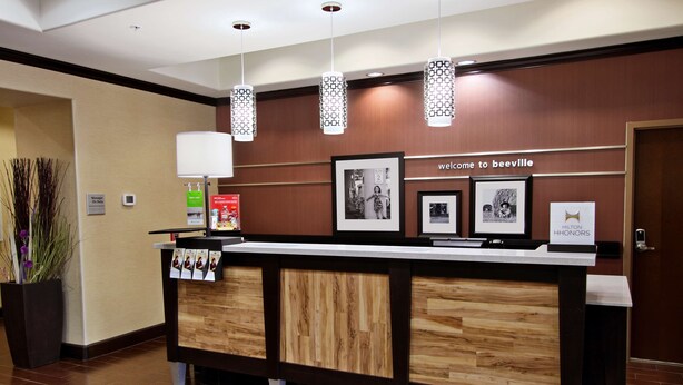 A hotel reception desk with a wooden finish, framed welcome signs, and a digital screen displaying the hotel name.