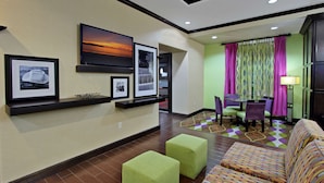 A hotel lobby with a reception desk, framed pictures on the wall, a seating area with a patterned sofa, and a dining area with a table and chairs.
