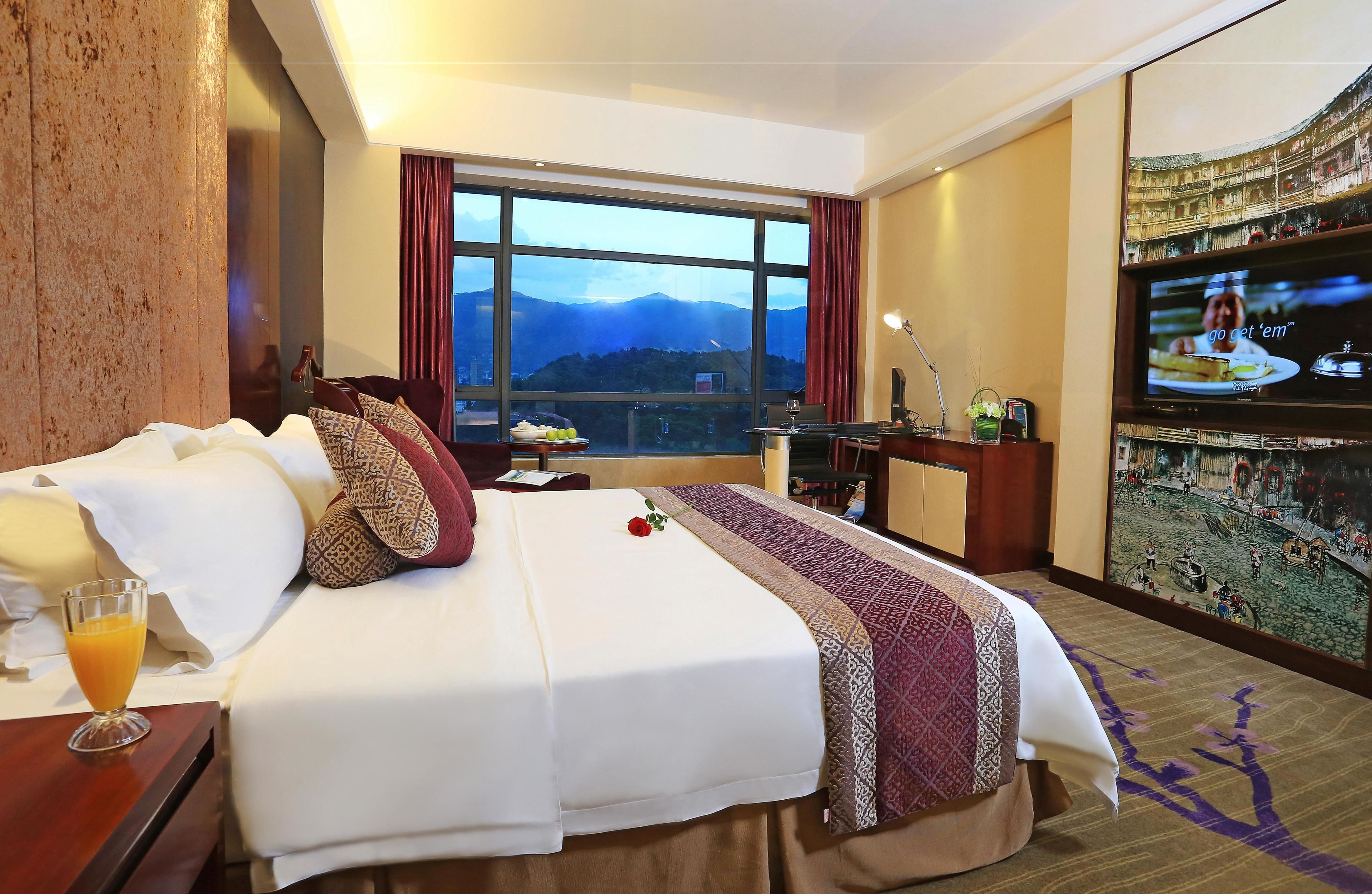 A hotel room with a large bed, a TV, a desk, and a view of mountains through the window.