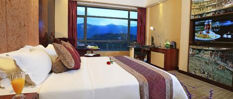 A hotel room with a large bed, a TV, a desk, and a view of mountains through the window.