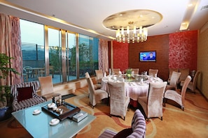 A modern dining area with a round table set for six, surrounded by patterned chairs. A television is mounted on the wall, and the room features large windows and a ceiling chandelier. - Ramada Longyan (Longyan)