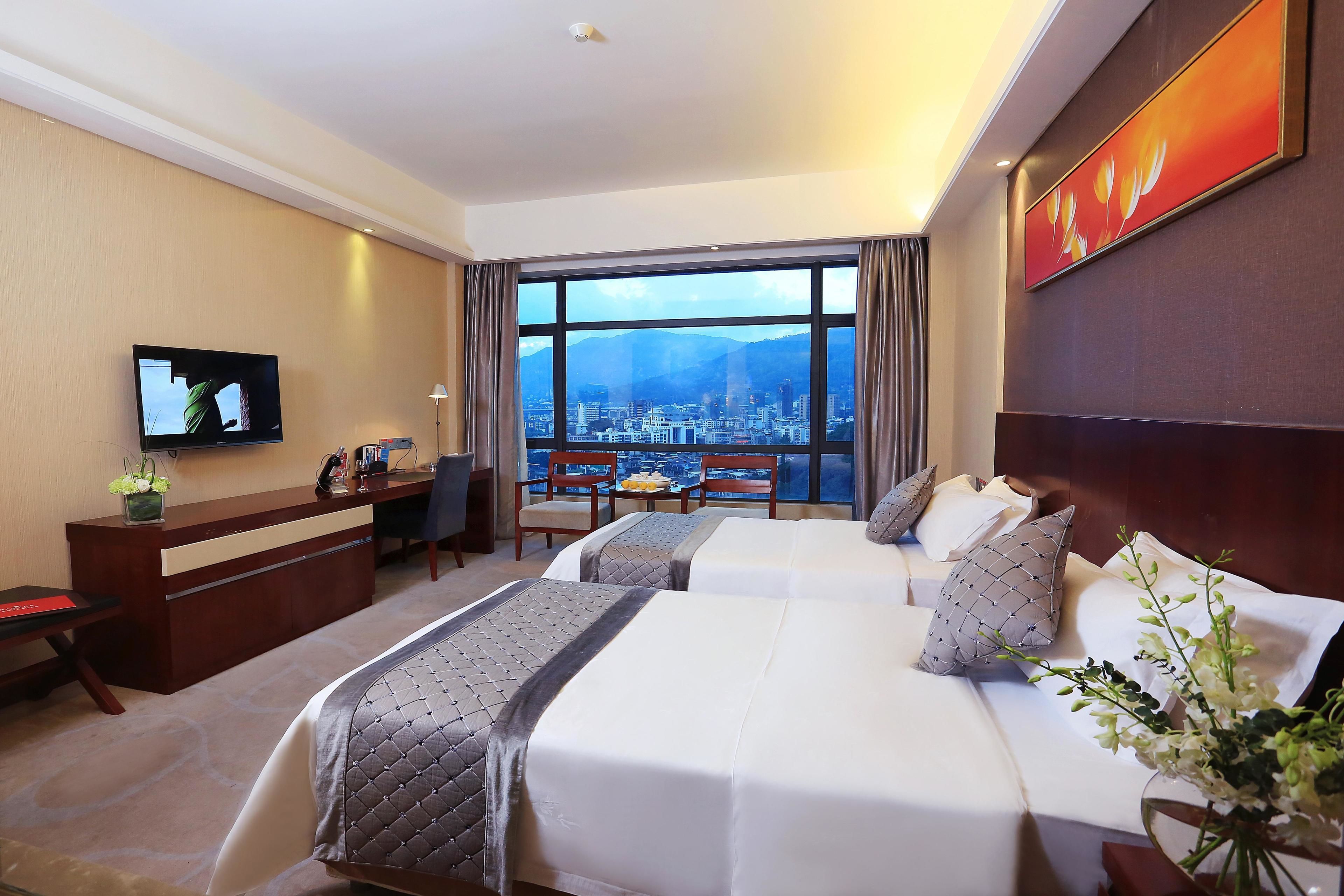 A hotel room with two beds, a desk, a TV, and a large window with a city view.