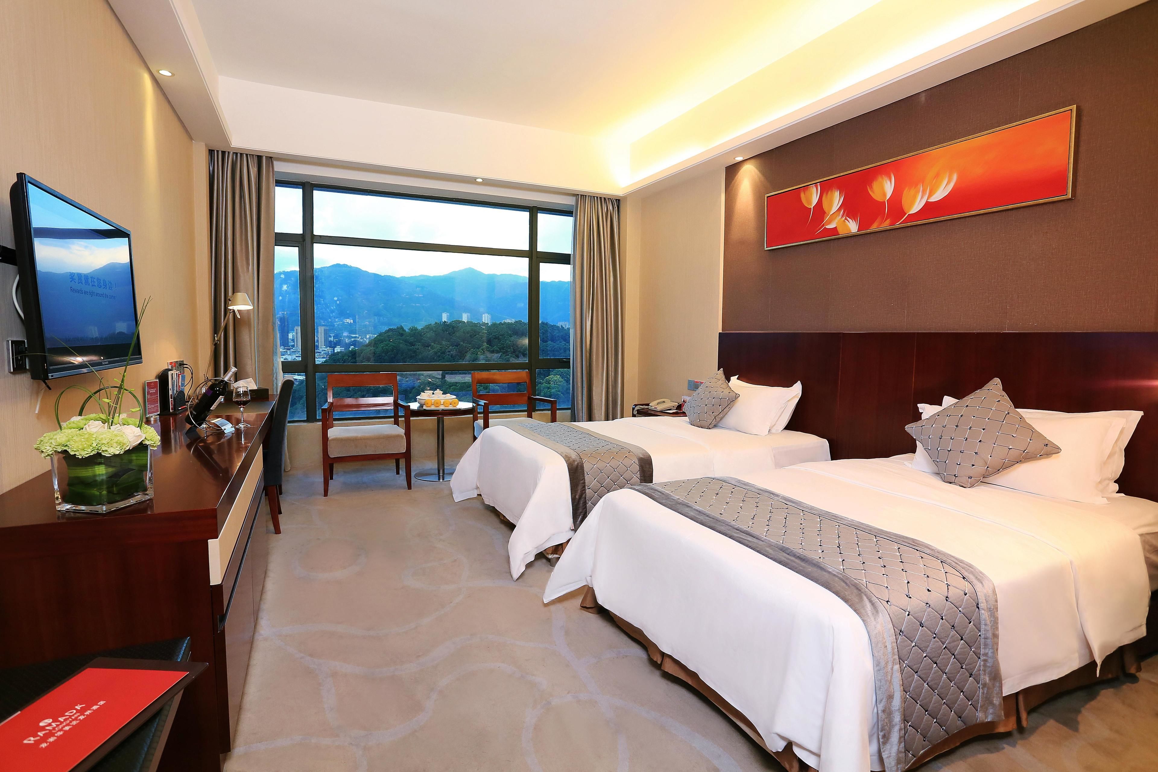 A hotel room with two beds, a desk, a TV, and a large window with a mountain view.
