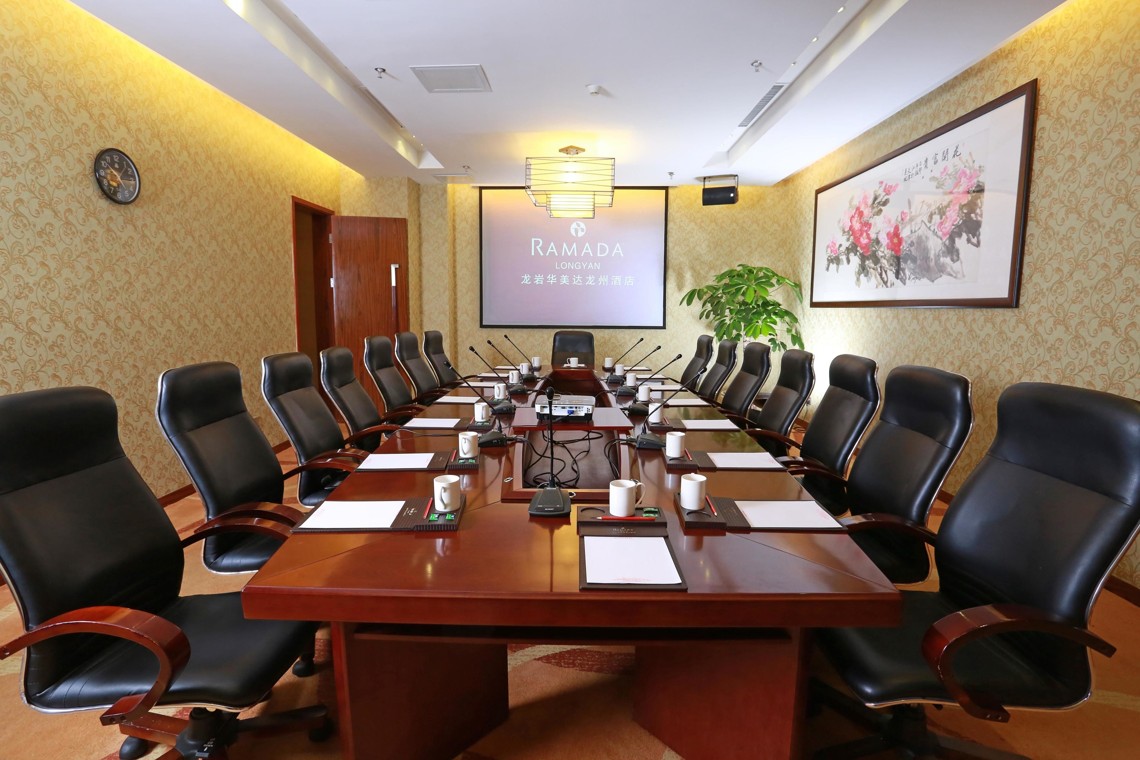 A conference room with a long table, black chairs, microphones, and a screen displaying "Ramada LONGYAN".