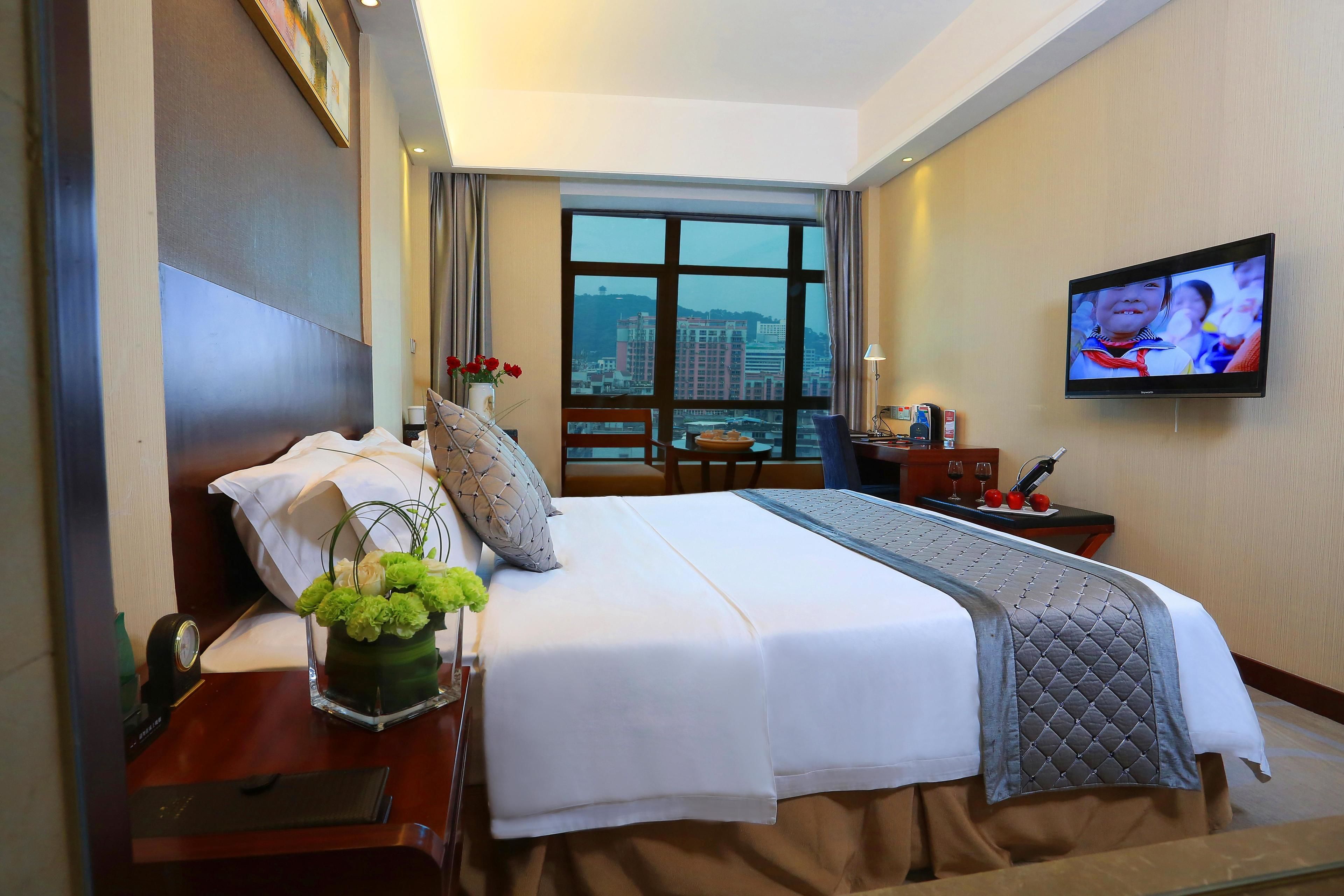 Una habitación de hotel con una cama grande, un televisor en la pared, un ventanal con vistas a la ciudad y un escritorio con un jarrón de flores.