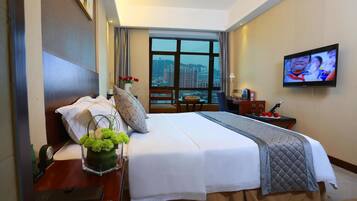 Una habitación de hotel con una cama grande, un televisor en la pared, un ventanal con vistas a la ciudad y un escritorio con un jarrón de flores.