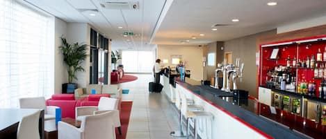 A modern bar area with a long counter, seating with red and white chairs, and a bar stocked with various bottles.