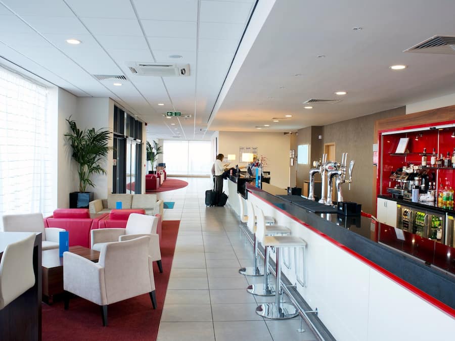 A modern bar area with a long counter, seating with red and white chairs, and a bar stocked with various bottles.