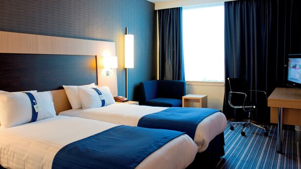 Quarto de hotel com duas camas, uma escrivaninha, uma cadeira, uma televisão e uma janela com cortinas.