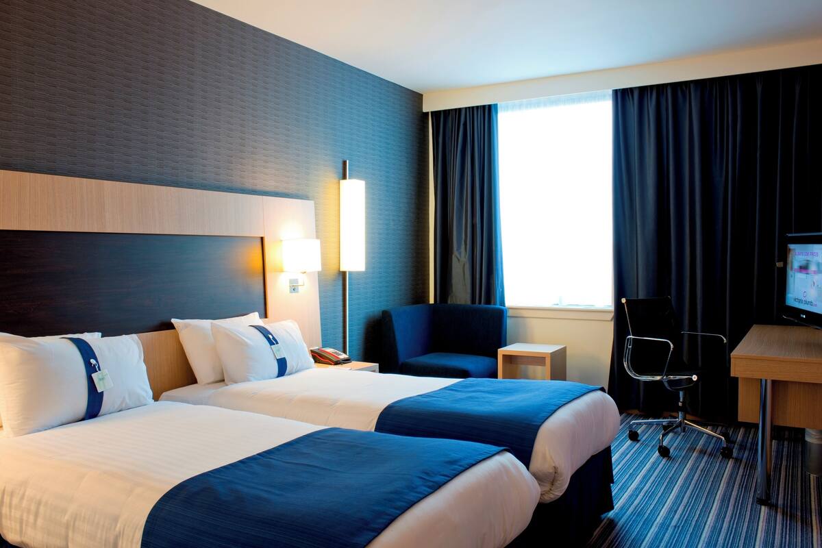 A hotel room with two beds, a desk, a chair, a TV, and a window with curtains.