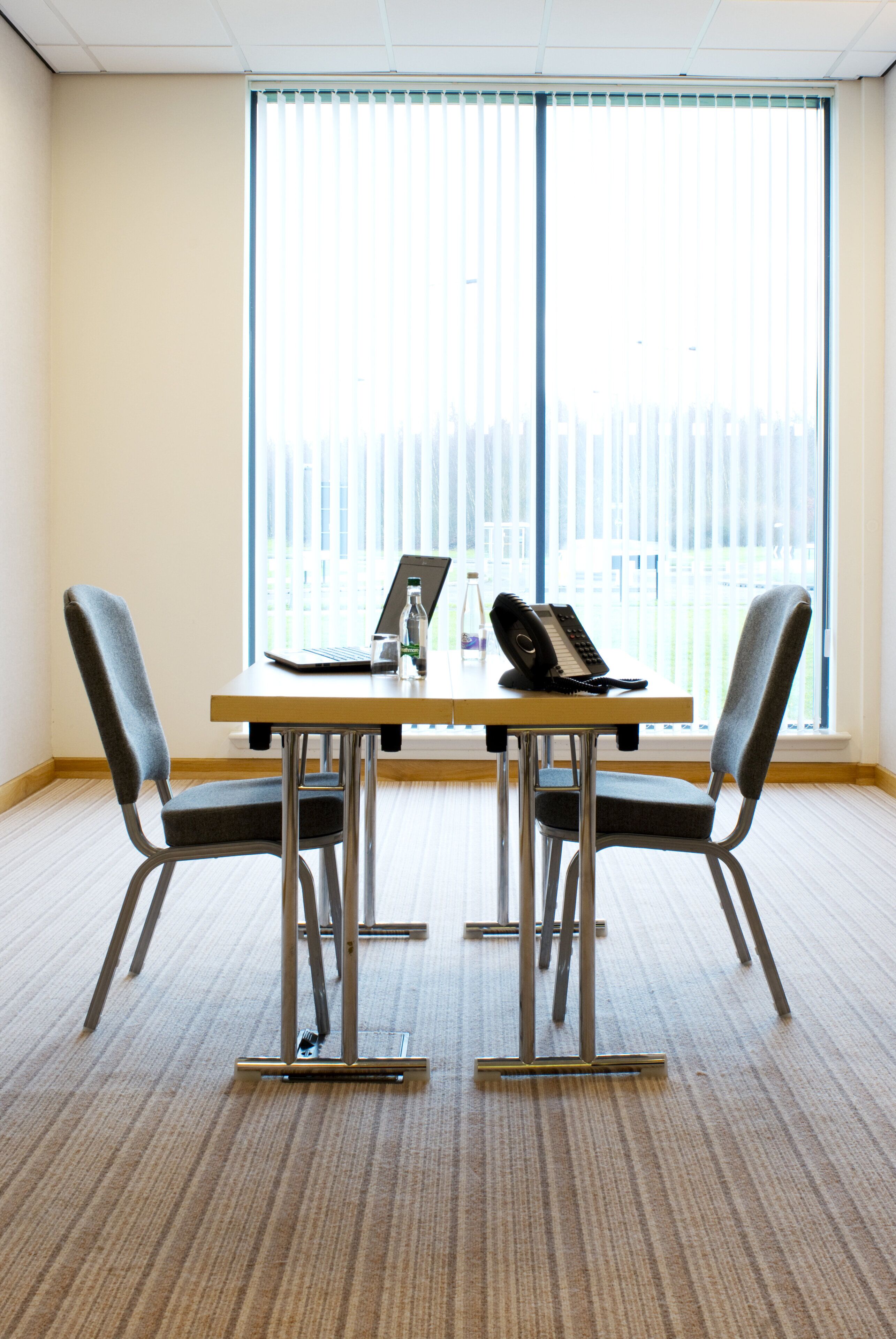A conference room with a table, two chairs, a laptop, a bottle, and a telephone.