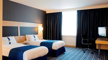 A hotel room with two beds, a desk with a chair, a television, and a large window with curtains.