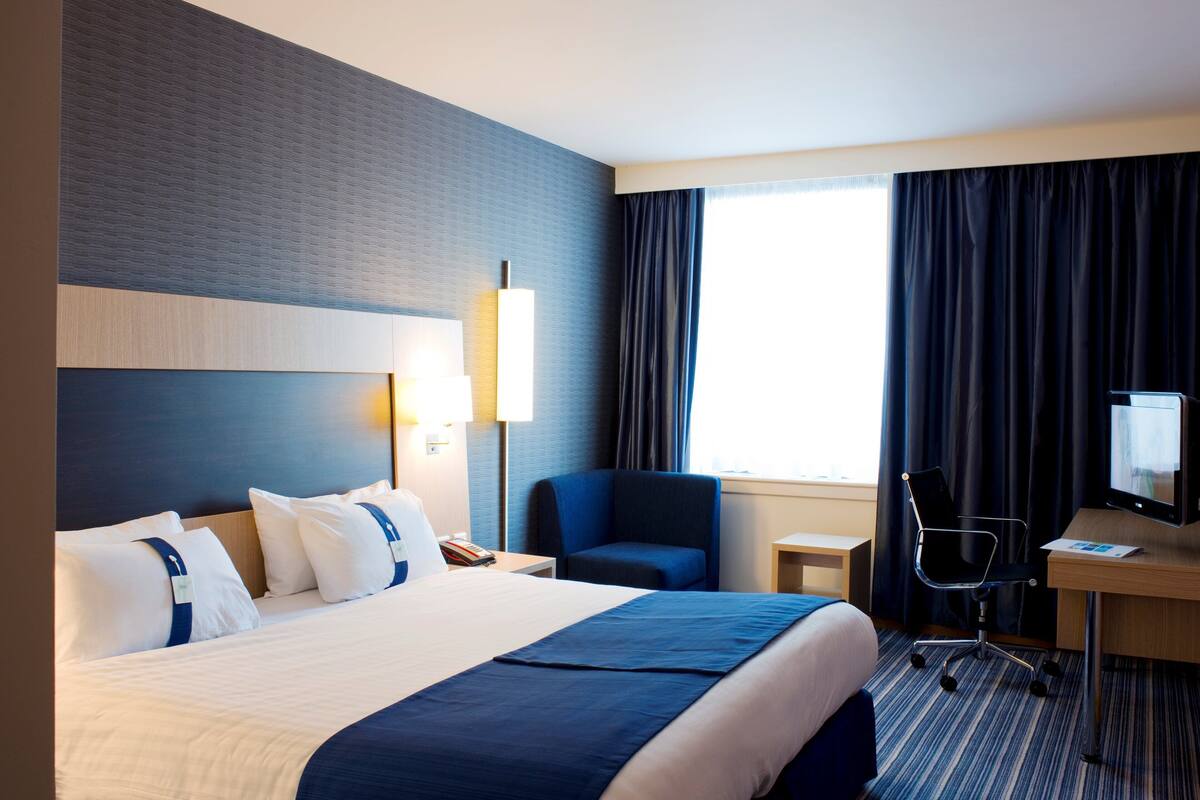 A hotel room with a large bed, a desk with a computer, a chair, and a window with curtains.