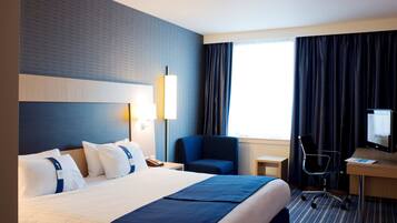 Ein Hotelzimmer mit einem großen Bett, einem Schreibtisch mit Computer, einem Stuhl und einem Fenster mit Vorhängen.