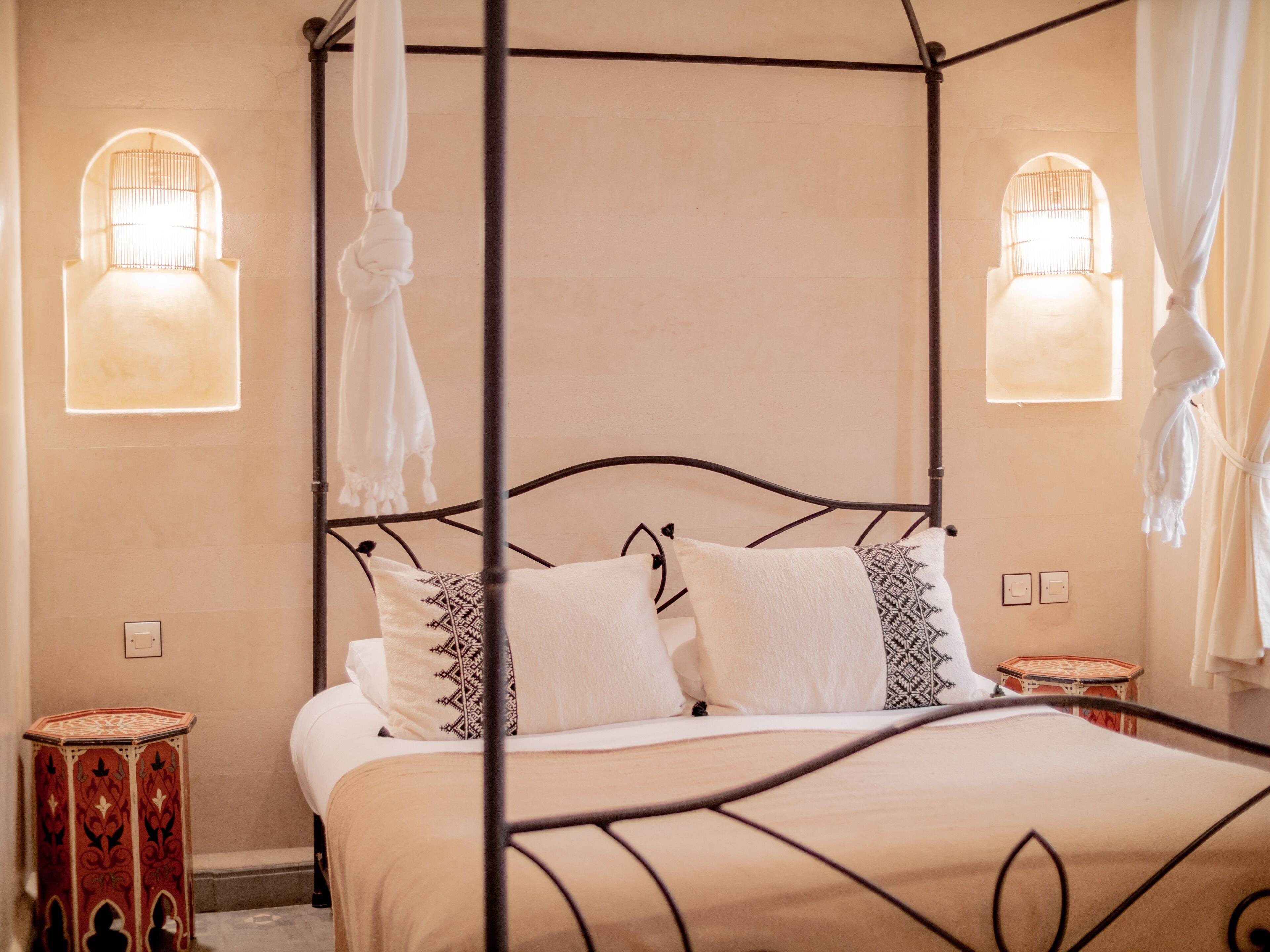 A four-poster bed with white linens and patterned pillows, a red and white decorative stool, and two wall-mounted lamps.