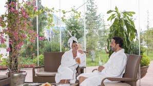 A couple in white bathrobes sitting on wicker chairs in a sunlit room with a glass table, surrounded by potted plants and a view of greenery. - NG Afyon (Afyonkarahisar)