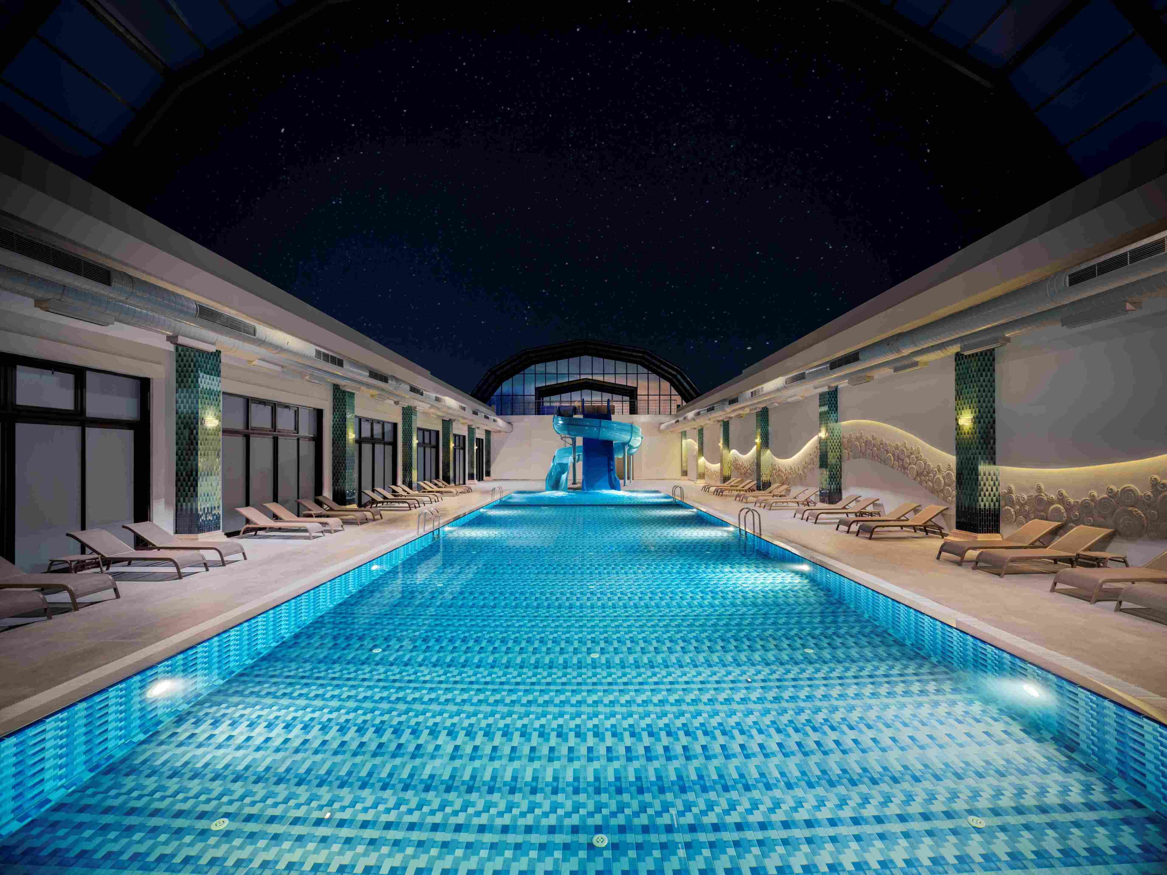A swimming pool with a water slide, lounge chairs, and a night sky view.