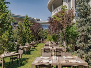 An outdoor dining area with tables and chairs, surrounded by trees and a building in the background. - NG Afyon (Afyonkarahisar)