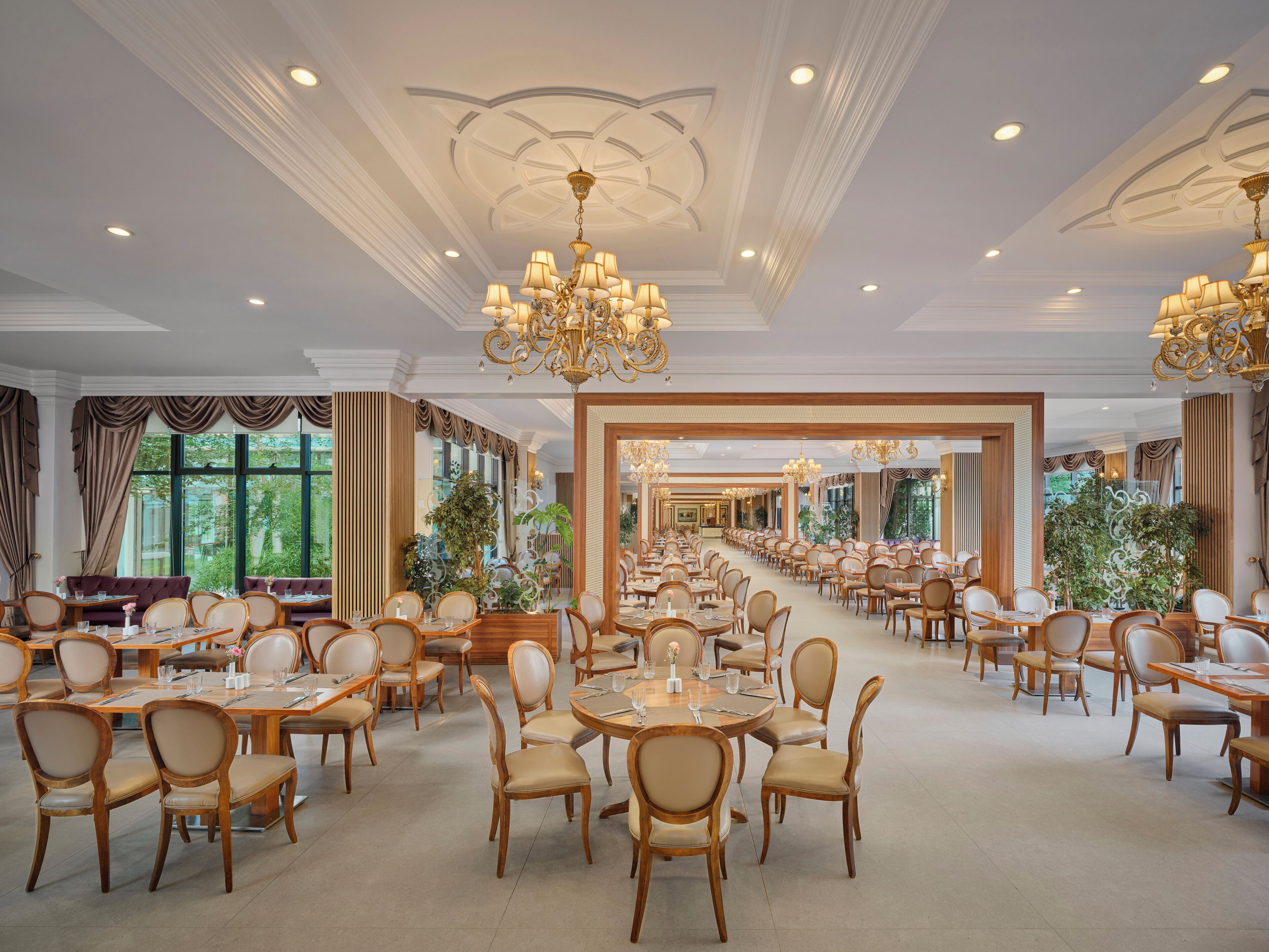 Un comedor amplio con varias mesas y sillas, candelabros elegantes y grandes ventanales con cortinas.