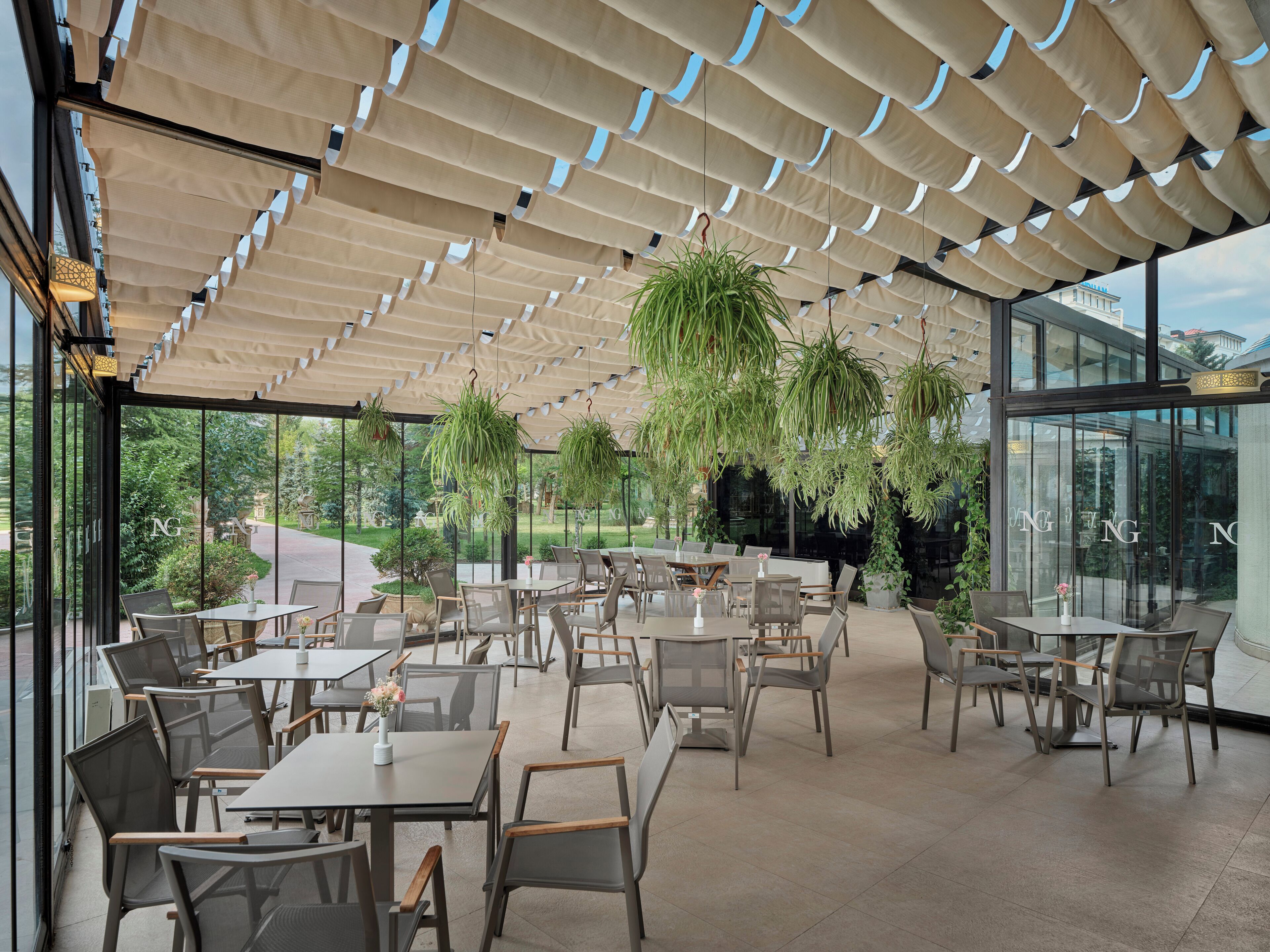 An outdoor dining area with tables and chairs, hanging plants, and a glass wall with the letters 'NG' visible.