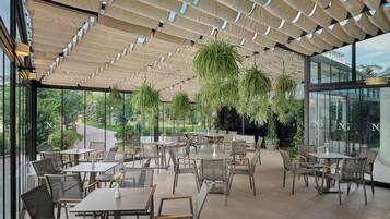 An outdoor dining area with tables and chairs, hanging plants, and a glass wall with the letters 'NG' visible.