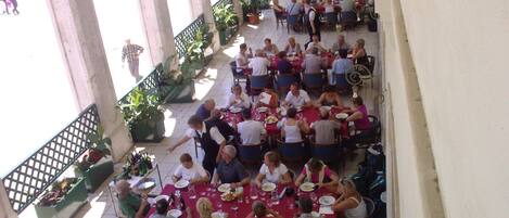 Un espacio comedor al aire libre con techo, varias mesas y sillas, rodeado de columnas y plantas en macetas.