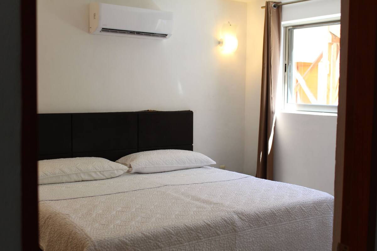 Une chambre à coucher avec un lit, du linge de lit blanc, une tête de lit sombre, un climatiseur, une fenêtre avec des rideaux et un luminaire fixé au mur.