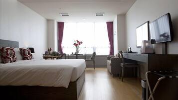A hotel room with a large bed, a desk with a chair, a TV mounted on the wall, and a window with curtains.