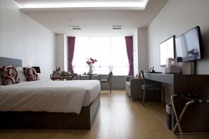 A hotel room with a large bed, a desk with a chair, a TV mounted on the wall, and a window with curtains.