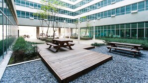 A modern building with a courtyard featuring wooden picnic tables and benches, surrounded by glass windows and greenery. - Hotel Hu (Incheon)