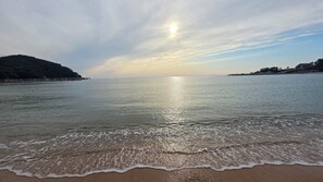 Una playa con olas suaves, cielo despejado y una lejana vista de un paisaje montañoso.