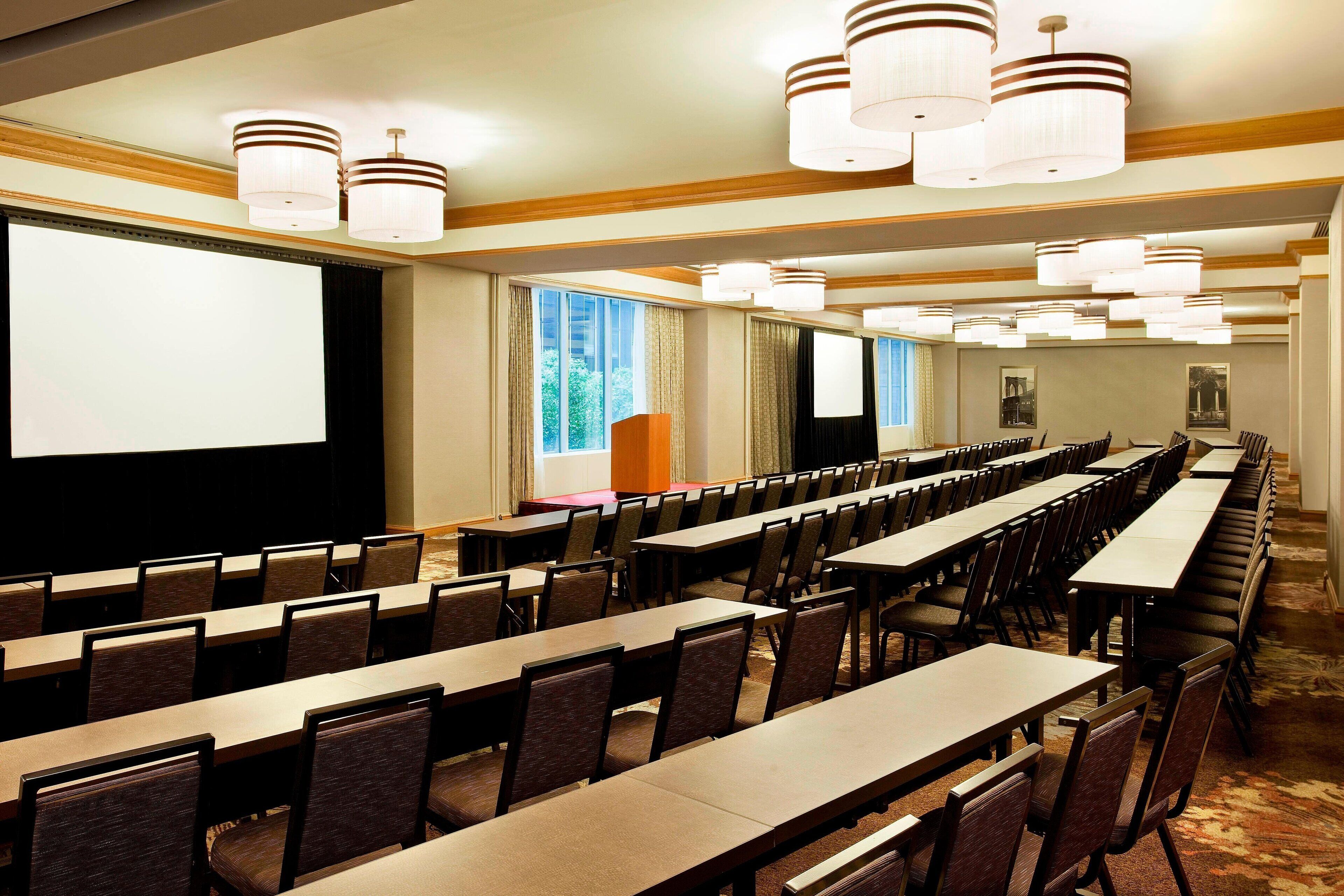 A conference room with rows of tables and chairs, a large screen, and multiple chandeliers.