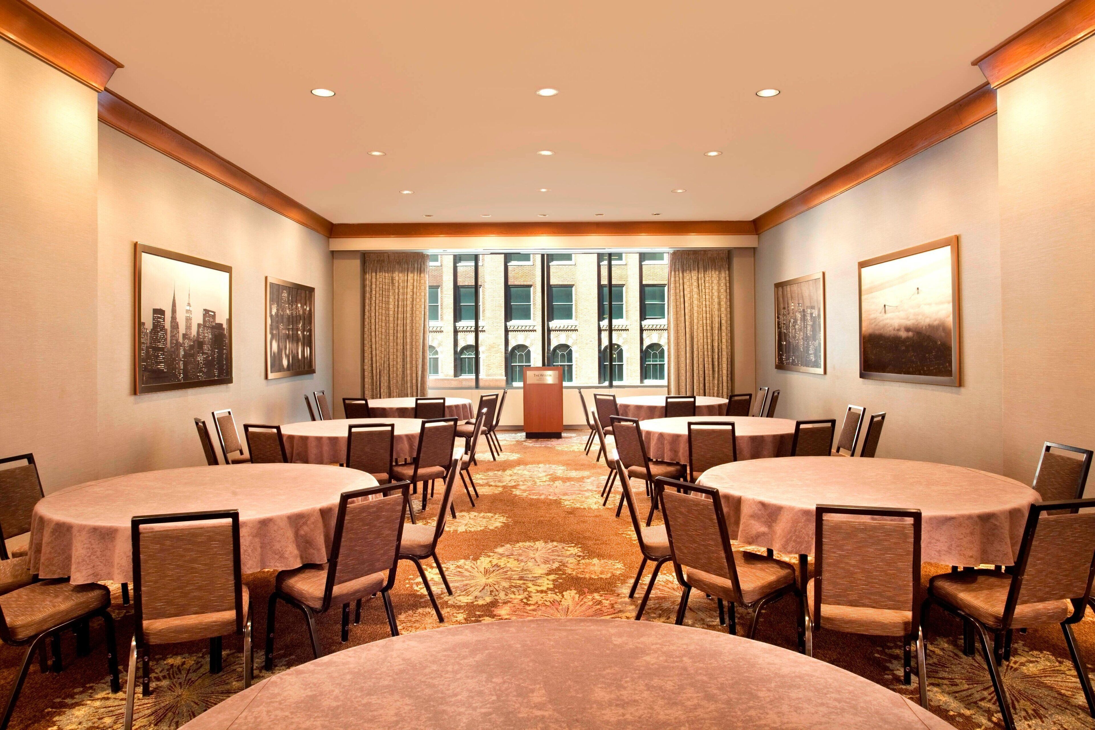 A conference room with multiple round tables and chairs, a podium, and large windows with a cityscape view.