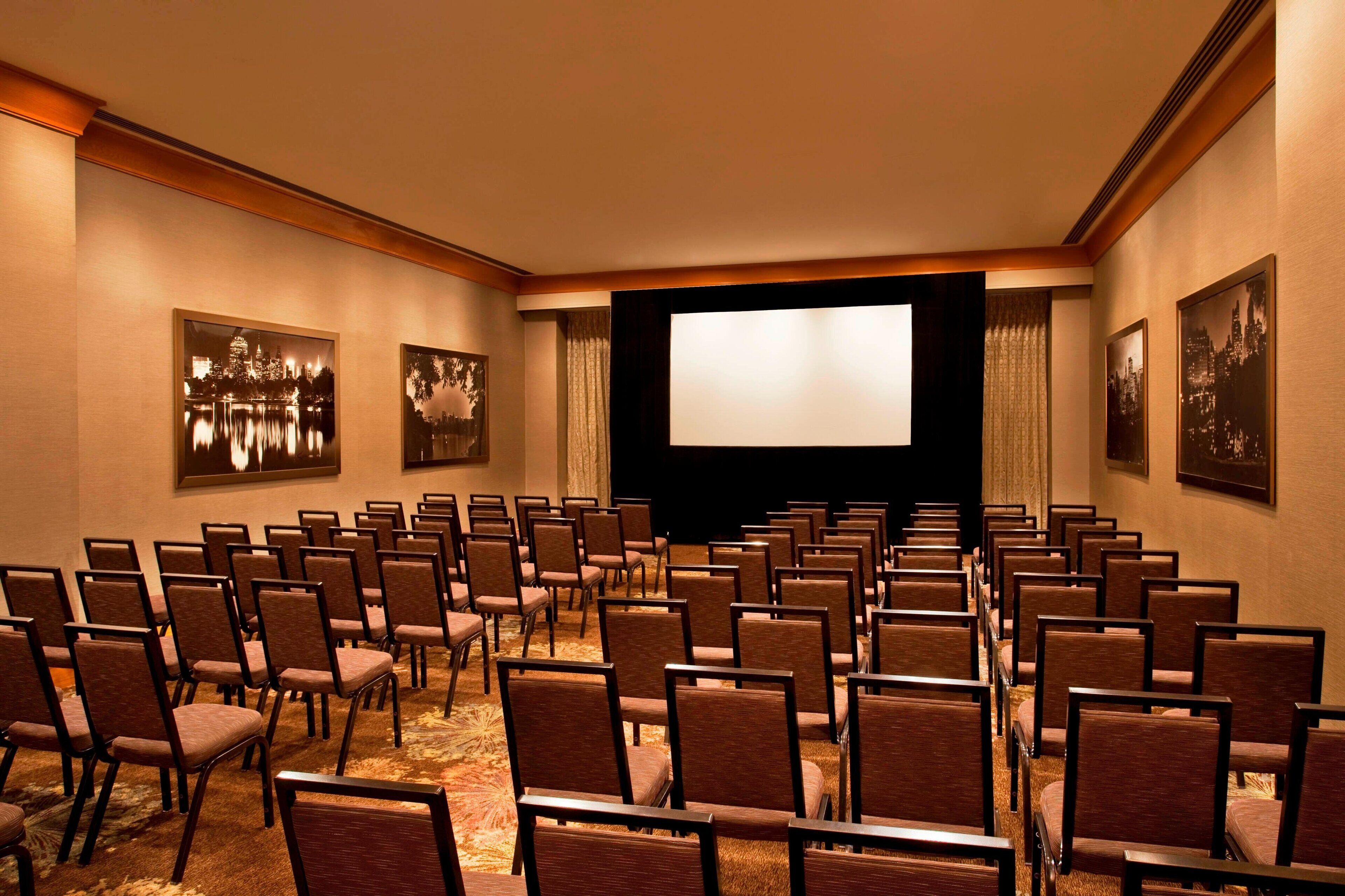 A conference room with rows of chairs facing a large screen, framed pictures on the walls, and a carpeted floor.