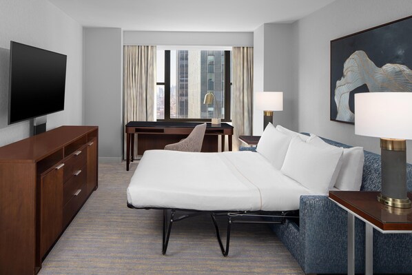 Habitación de hotel con una cama grande, un escritorio, un televisor y vistas a la ciudad desde la ventana.