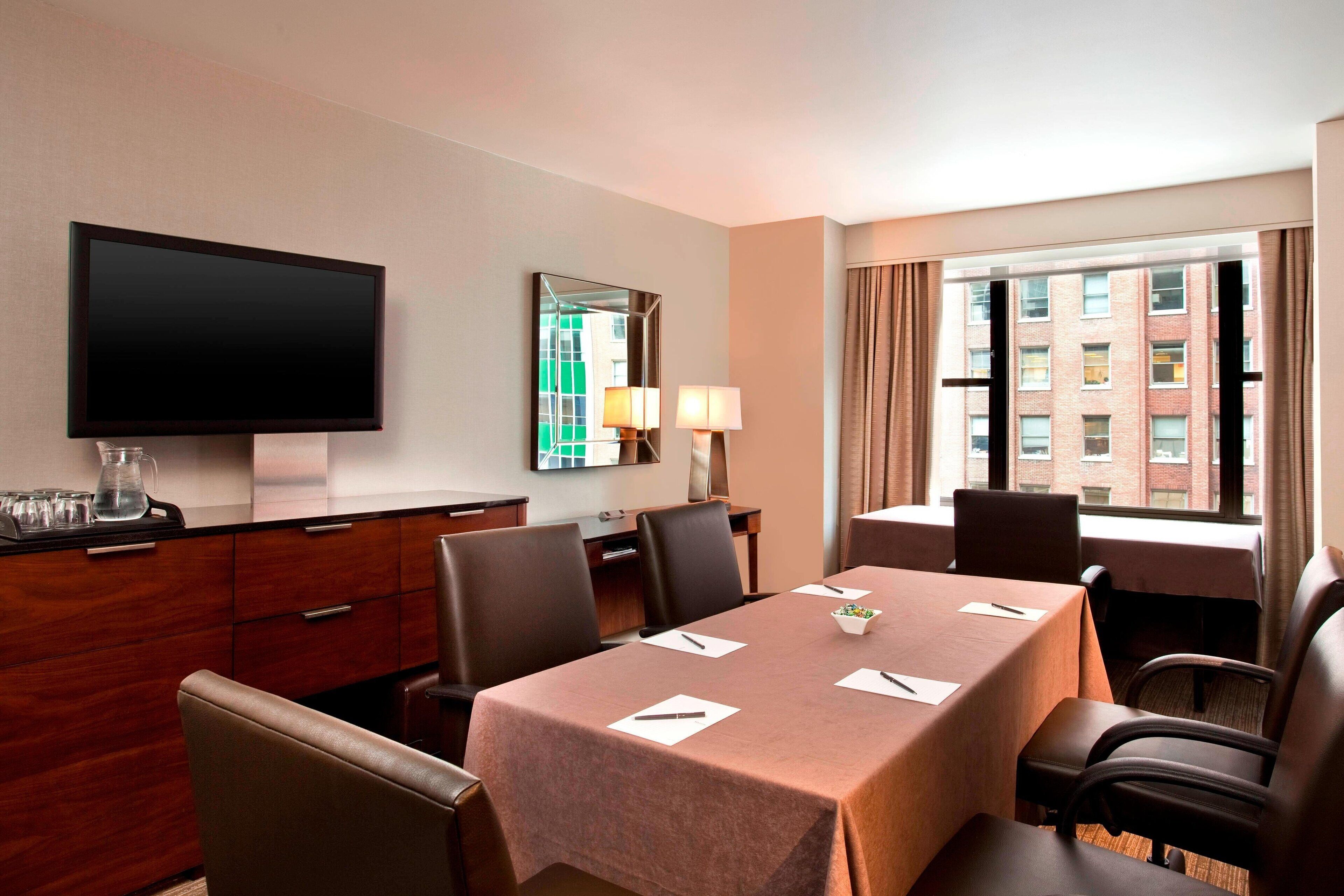 A hotel conference room with a large table, chairs, a television, a mirror, and a window with curtains.