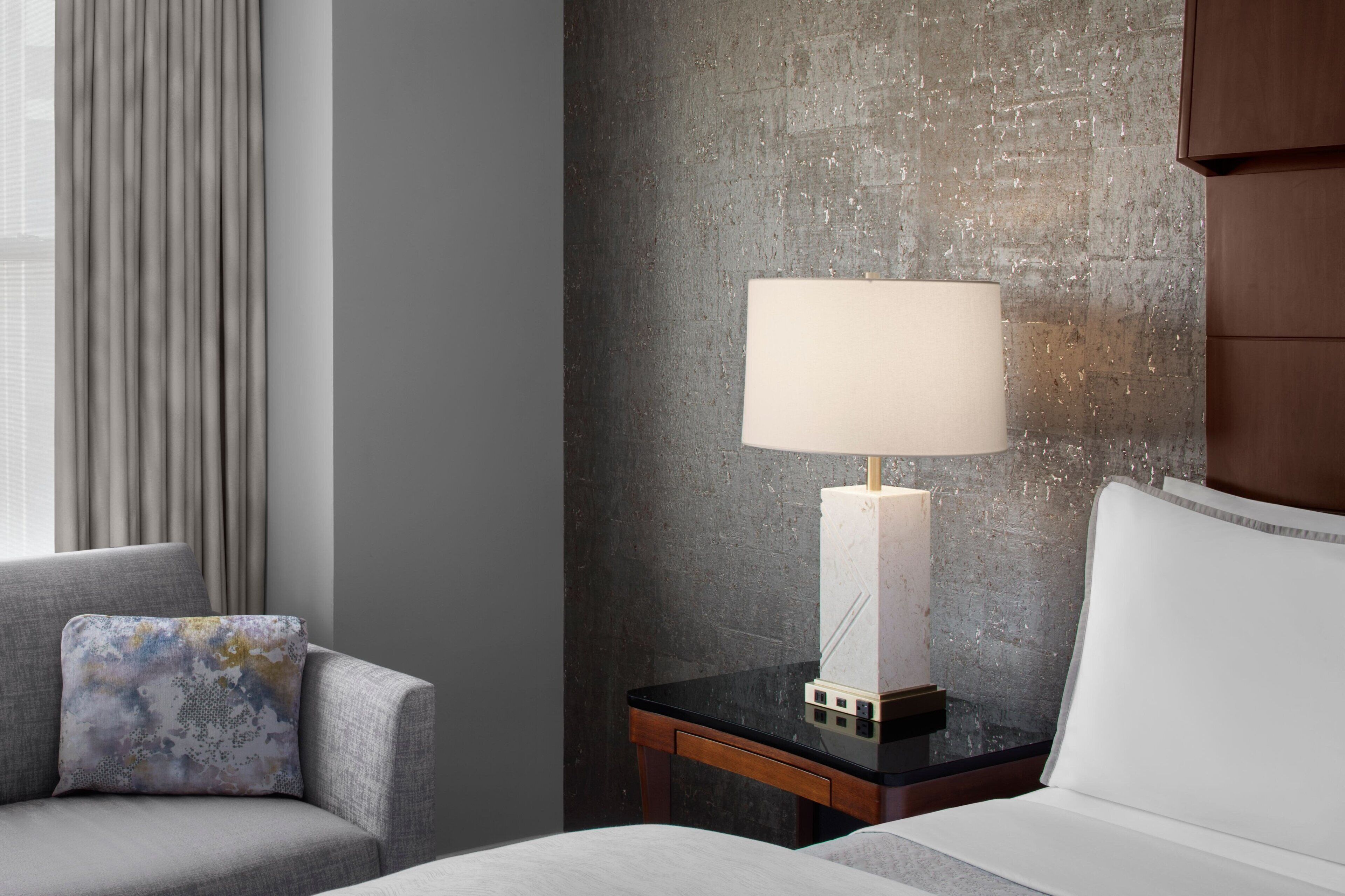 A hotel room with a bed, a nightstand with a lamp, a chair, and a view of the outdoors.