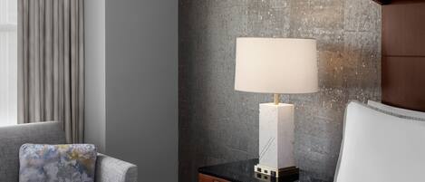 Habitación de hotel con cama, mesita de noche con lámpara, una silla y vistas al exterior.