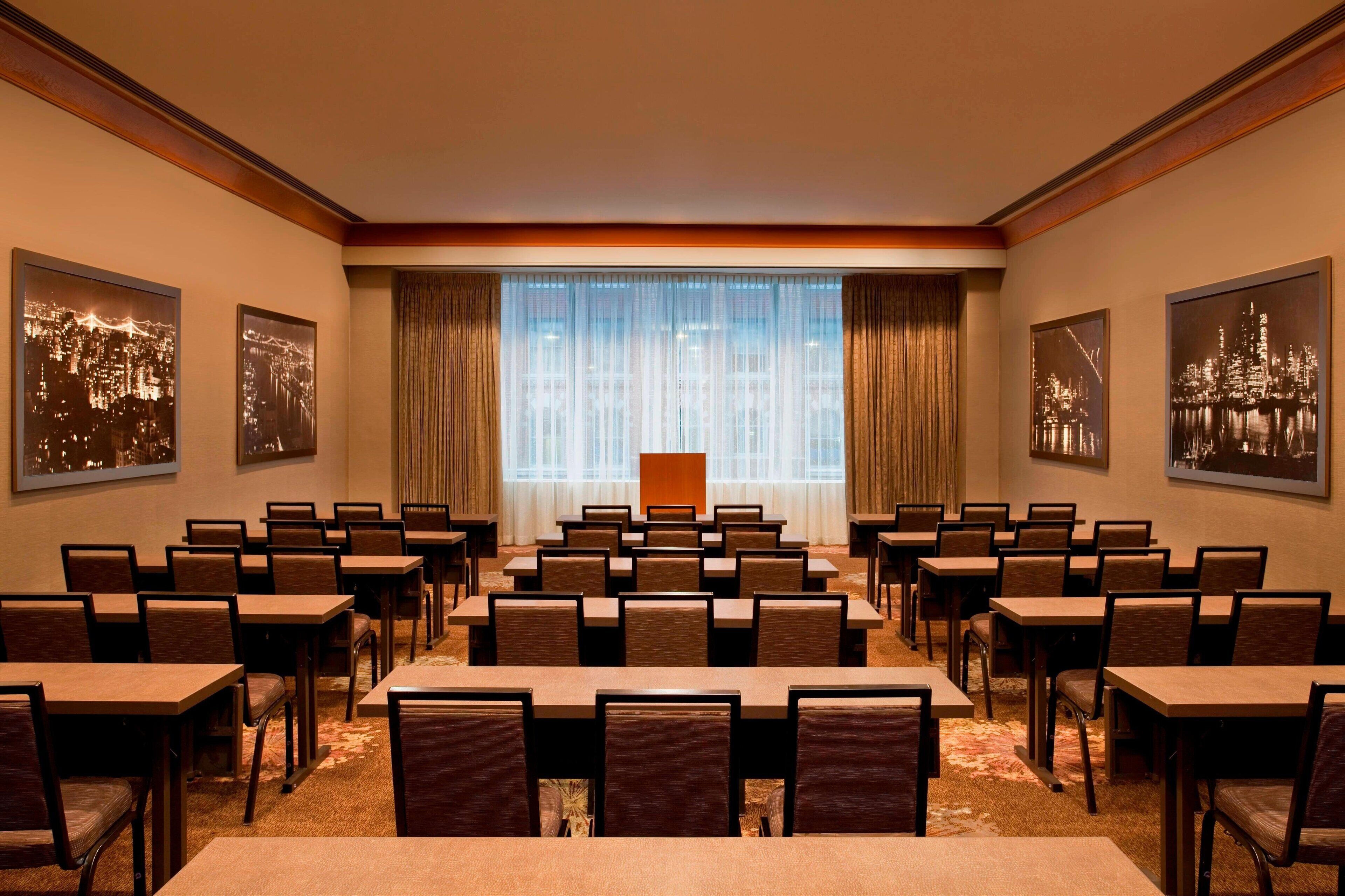 Un salón de conferencias con filas de mesas y sillas, ventanas amplias con cortinas y fotografías enmarcadas de la ciudad.