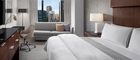 A hotel room with a bed, a sofa, a desk, and a view of the cityscape.