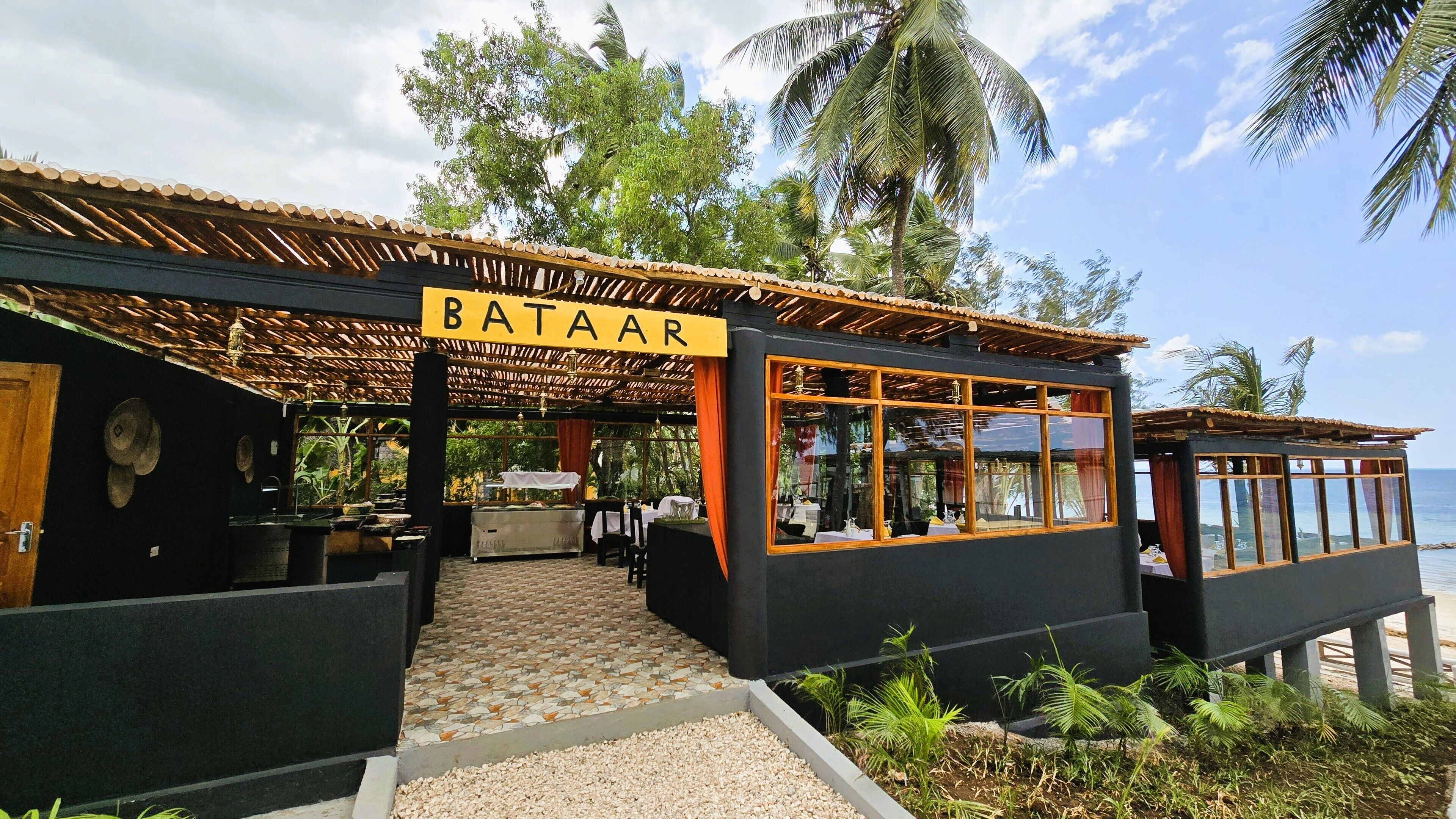 Ein BATAAR Restaurant mit tropischer Atmosphäre, das eine überdachte Außenbereich-Sitzgelegenheit, einen gefliesten Weg und einen Blick auf den Ozean bietet.