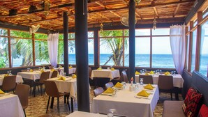 A restaurant with tables set for dining, large windows with a view of the ocean, and a thatched roof. - Paradise Beach Resort (Uroa)