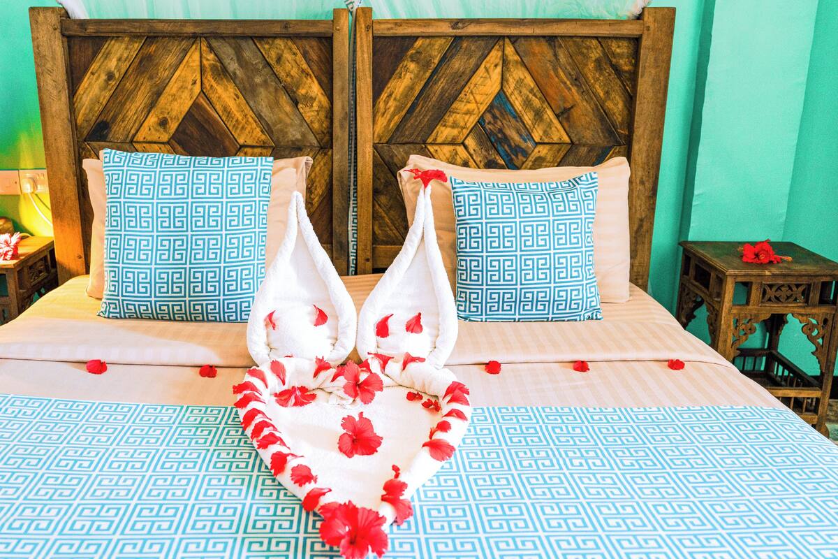 Uma cama com colcha estampada, duas almofadas com padrão geométrico e um arranjo decorativo de flores vermelhas sobre a cama.