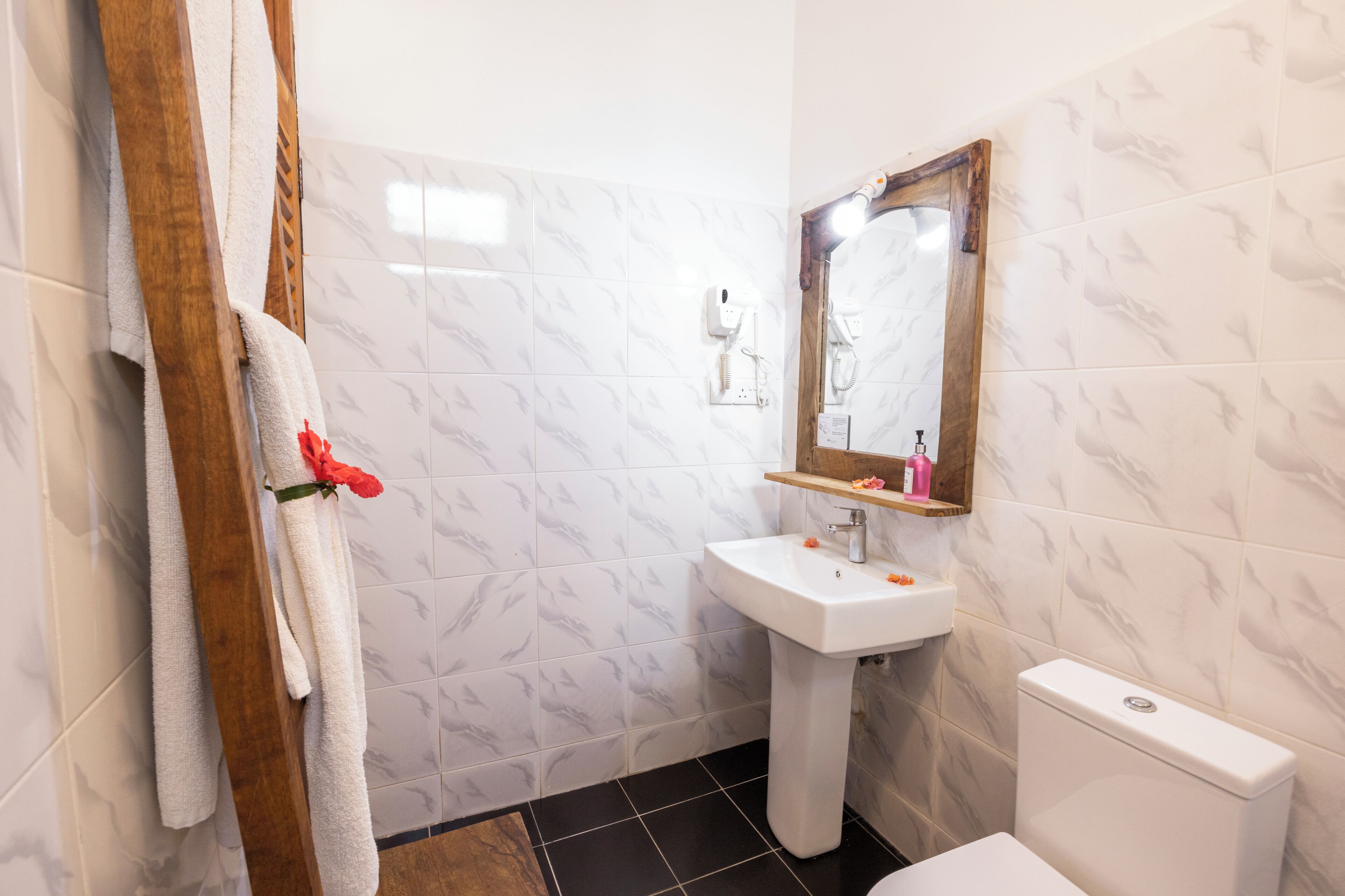 A bathroom with a white marble wall, a wooden towel rack with a white towel, a white pedestal sink, a mirror, and a white toilet.