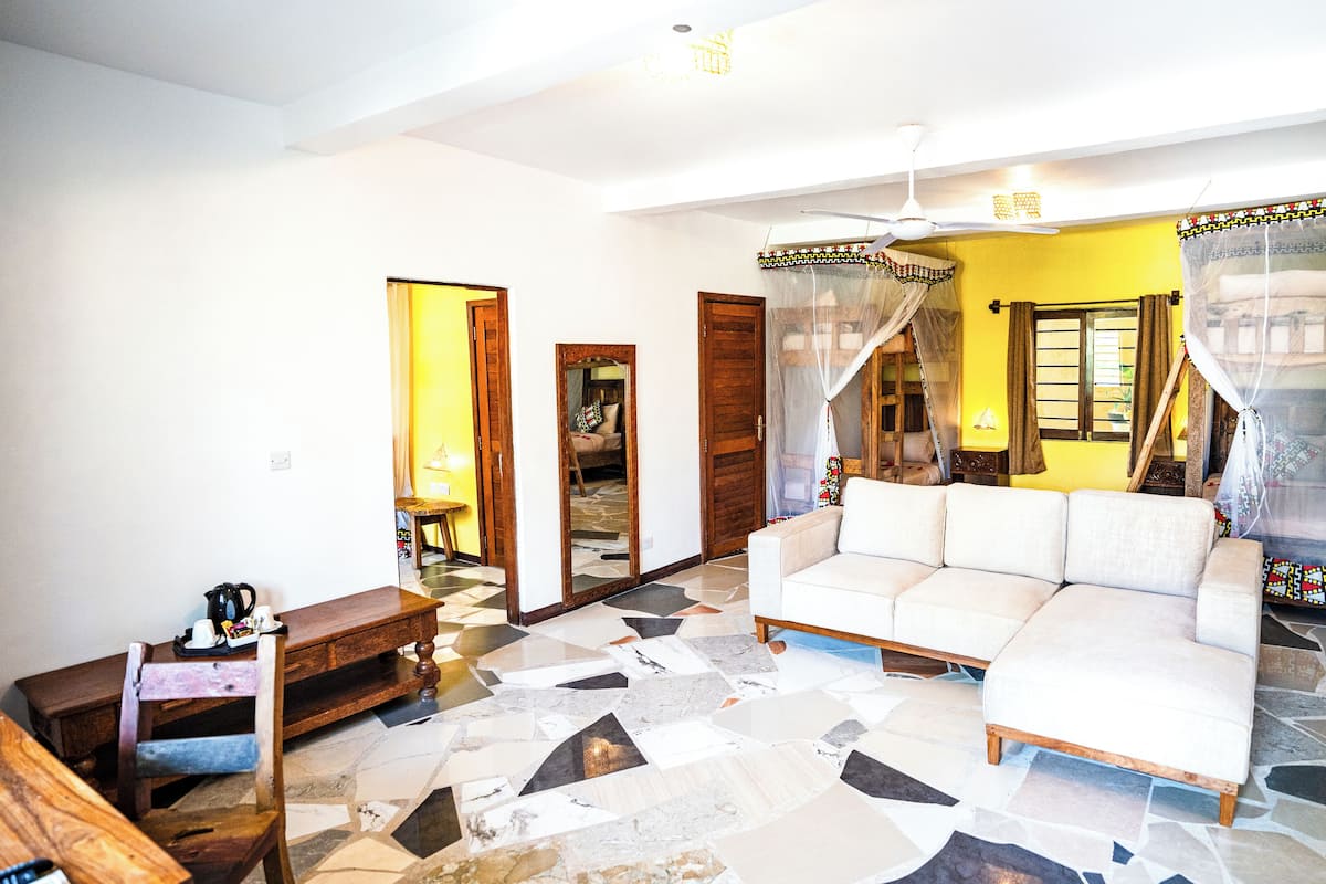 Uma sala de estar com sofá, mesa de centro e televisão. Há um espelho, uma porta e uma janela com cortinas.