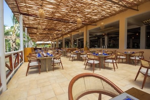 An outdoor dining area with tables and chairs, a thatched roof, and a view of the outdoors.