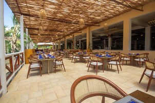 Un área de comedor al aire libre con mesas y sillas, techo de paja y vistas del exterior.