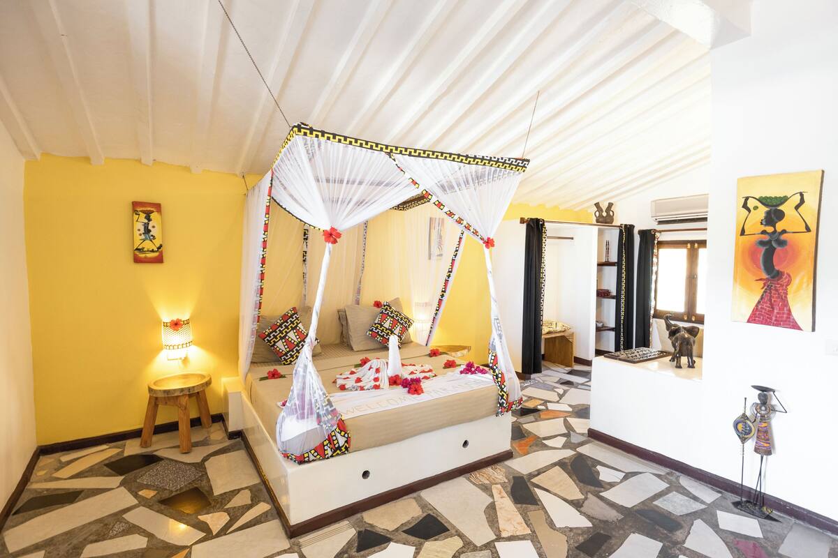 Quarto com cama de dossel, um banquinho, luminária na parede e um quadro.