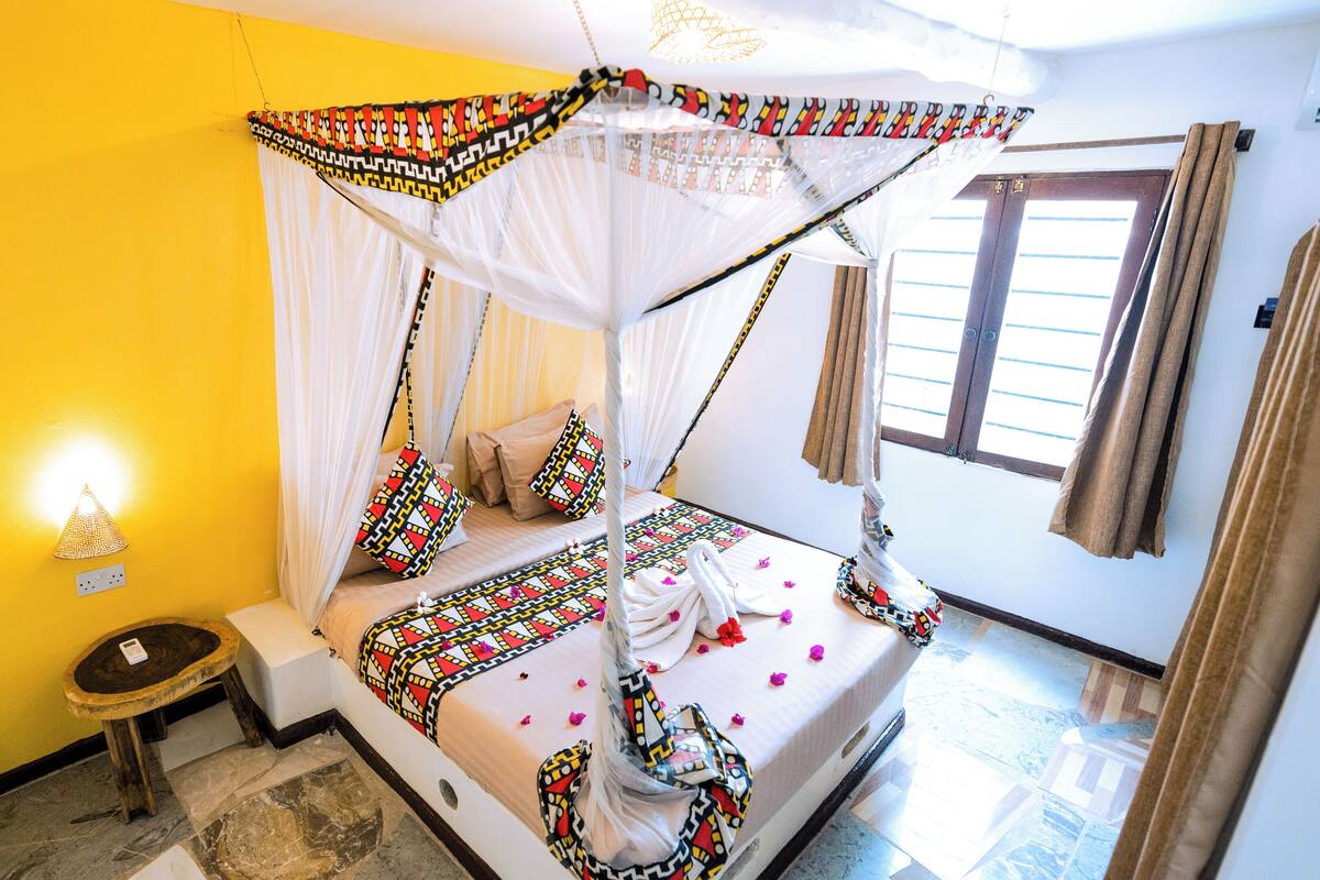 Quarto com uma cama, uma mesinha, uma luminária na parede e uma janela com cortinas.
