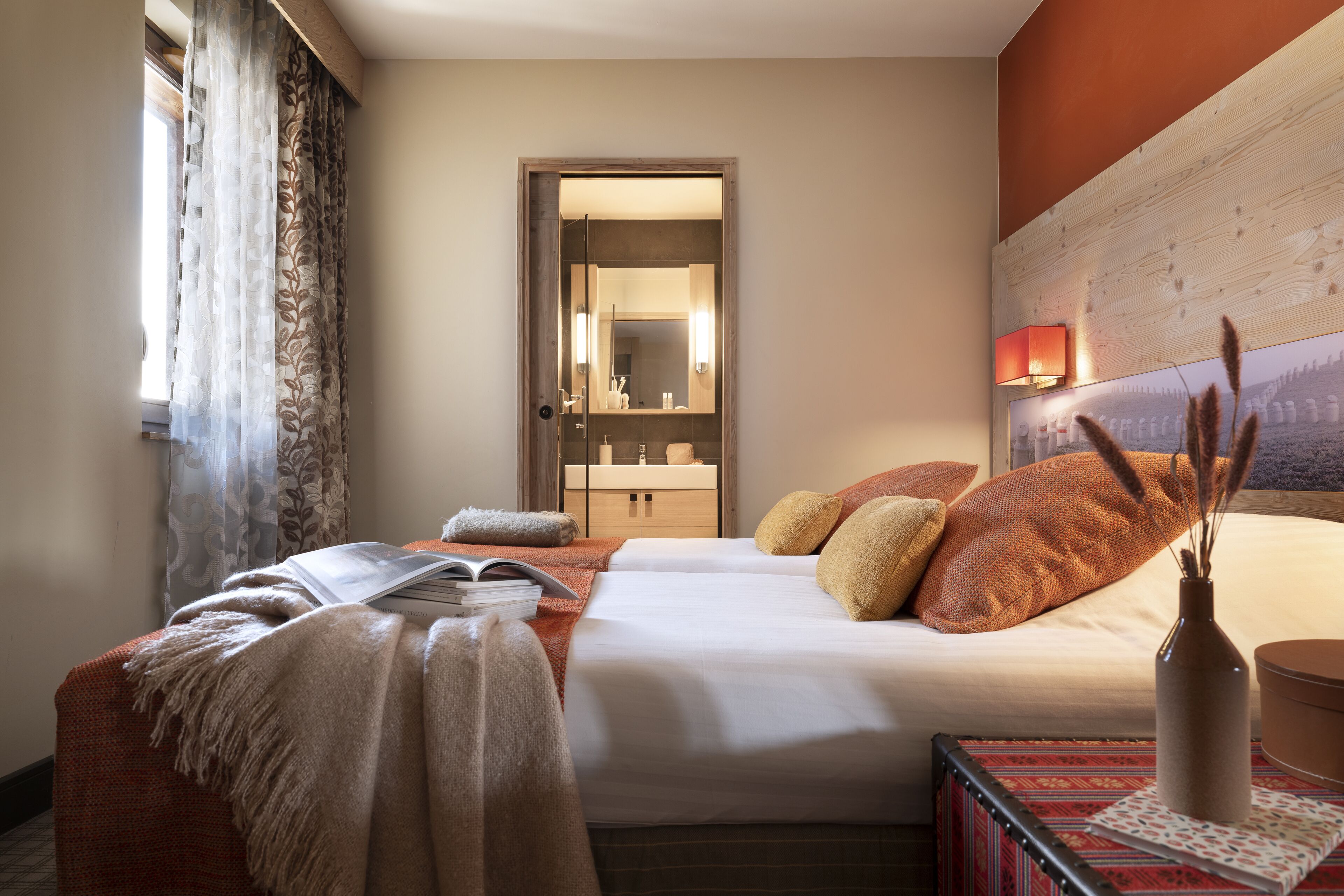 A hotel room with a bed, a bedside table with a book and a vase, a window with curtains, and a bathroom visible through an open door.